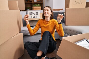 Poster - Young caucasian woman sitting on the floor at new home holding new home banner smiling and laughing hard out loud because funny crazy joke.