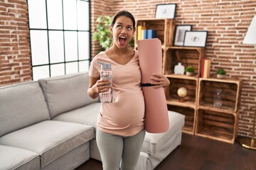 Sticker - Young pregnant woman holding yoga mat at home angry and mad screaming frustrated and furious, shouting with anger looking up.