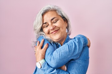 Poster - Middle age woman with grey hair standing over pink background hugging oneself happy and positive, smiling confident. self love and self care