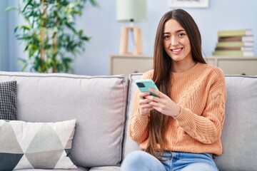 Wall Mural - Young beautiful hispanic woman using smartphone sitting on sofa at home