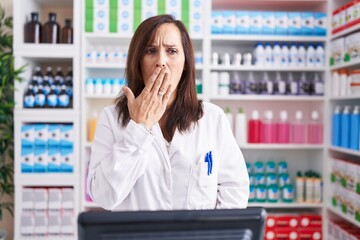 Poster - Middle age brunette woman working at pharmacy drugstore bored yawning tired covering mouth with hand. restless and sleepiness.