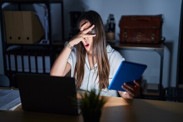 Poster - Young brunette woman working at the office at night peeking in shock covering face and eyes with hand, looking through fingers with embarrassed expression.