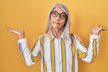 Poster - Middle age woman with grey hair standing over yellow background wearing glasses clueless and confused expression with arms and hands raised. doubt concept.