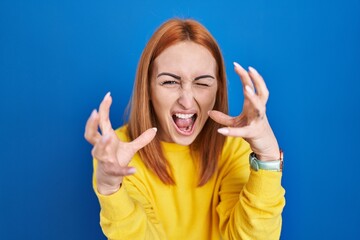 Poster - Young woman standing over blue background shouting frustrated with rage, hands trying to strangle, yelling mad