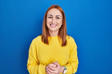 Wall Mural - Young woman standing over blue background with hands together and crossed fingers smiling relaxed and cheerful. success and optimistic