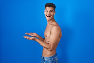 Canvas Print - Young hispanic man standing shirtless over blue background pointing aside with hands open palms showing copy space, presenting advertisement smiling excited happy