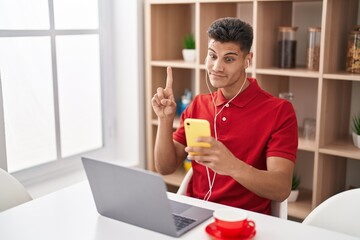 Canvas Print - Young hispanic man using laptop and doing video call with smartphone smiling with an idea or question pointing finger with happy face, number one