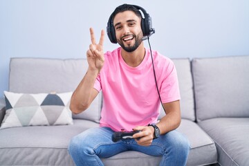 Wall Mural - Hispanic young man playing video game holding controller sitting on the sofa smiling with happy face winking at the camera doing victory sign with fingers. number two.