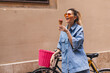 Happy laughing blonde girl with high ponytail in denim shirt and short , top outfit wear orange sunglasses and hold pink ice cream on a summer day next to a bright bike fixed. Street style, travel.
