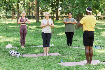 Wall Mural - Full length portrait of senior women enjoying yoga outdoors in park with male trainer assisting and demonstrating breathing and balance exercise