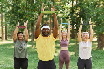 Wall Mural - Portrait of male sports instructor in park with senior women training with rubber bands in background