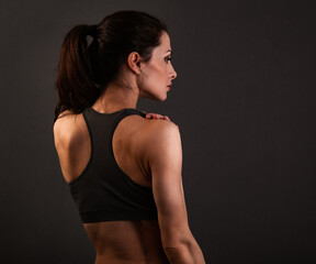 Wall Mural - Serious female sporty muscular with ponytail doing stretching workout the shoulders, blades and arms in sport bra, standing on dark grey background with empty copy space. Back view.