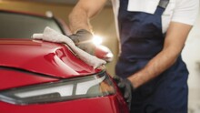 Low Angle View Of Male Professional Car Wash Worker In Black Rubber Gloves, Holding The Gray Microfiber And Polishing The Car Hood And Headlight Of Luxury Red Car. Car Wash Service