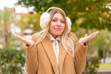 Wall Mural - Young pretty blonde woman wearing winter muffs at outdoors having doubts while raising hands