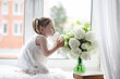 © alexkich - A little girl is sitting on the windowsill. A bouquet of flowers in a vase by the window and a girl sniffing flowers. A little princess in a white dress with a bouquet of white flowers by the window.