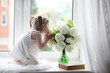 © alexkich - A little girl is sitting on the windowsill. A bouquet of flowers in a vase by the window and a girl sniffing flowers. A little princess in a white dress with a bouquet of white flowers by the window.