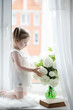© alexkich - A little girl is sitting on the windowsill. A bouquet of flowers in a vase by the window and a girl sniffing flowers. A little princess in a white dress with a bouquet of white flowers by the window.