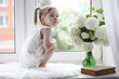 © alexkich - A little girl is sitting on the windowsill. A bouquet of flowers in a vase by the window and a girl sniffing flowers. A little princess in a white dress with a bouquet of white flowers by the window.