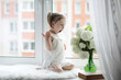 © alexkich - A little girl is sitting on the windowsill. A bouquet of flowers in a vase by the window and a girl sniffing flowers. A little princess in a white dress with a bouquet of white flowers by the window.
