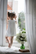 © alexkich - A little girl is sitting on the windowsill. A bouquet of flowers in a vase by the window and a girl sniffing flowers. A little princess in a white dress with a bouquet of white flowers by the window.