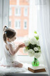 © alexkich - A little girl is sitting on the windowsill. A bouquet of flowers in a vase by the window and a girl sniffing flowers. A little princess in a white dress with a bouquet of white flowers by the window.