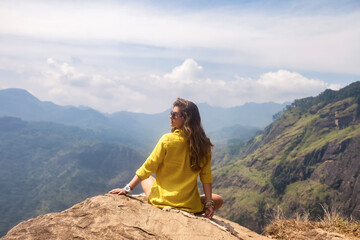 Rear view pretty woman sitting on rock in front of Sri Lankan mountains in nature background. Cute lady posing from behind enjoying at tropical journey, Ella, Sri Lanka. Copy advertising text space