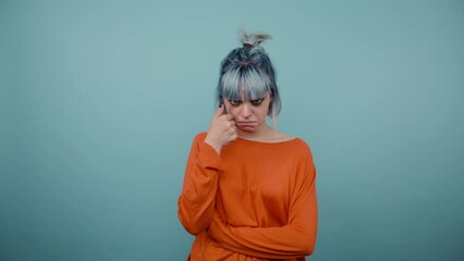 Wall Mural - Sad hipster girl with dyed turquoise hair shows a gesture of tears standing on a blue background and looking at the camera.