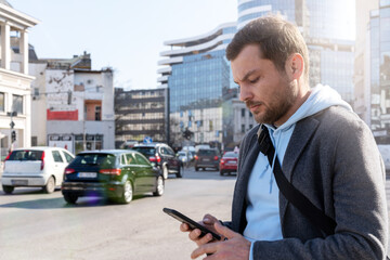 Man standing near the road in city and looking at screen of his smartphone, person using transportation mobile app.