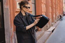 Elegant Young Woman Looking In Her Black Leather Bag Her Phone Or Purse. Business Style Woman Wear Grey Blazer, Black Eyeglasses And Bag On The Street. Street Style, Fashion Outfit.