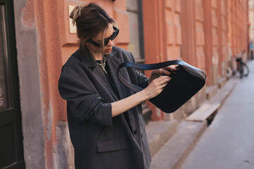 Wall Mural - Elegant young woman looking in her black leather bag her phone or purse. Business style woman wear grey blazer, black eyeglasses and bag on the street. Street style, fashion outfit.