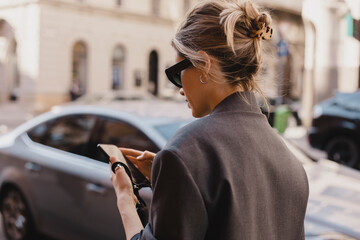 Wall Mural - Photo of young woman reading messages on a smartphone while standing on urban background. Blonde caucasian female wear is surfing the web on a mobile phone while walking the city. Focus on hair.