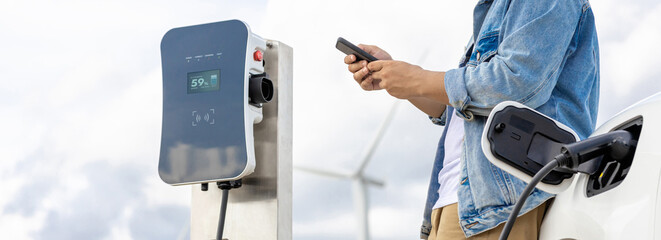 Wall Mural - Progressive man with his electric car, EV car recharging energy from charging station on green field with wind turbine as concept of future sustainable energy. Electric vehicle with energy generator.