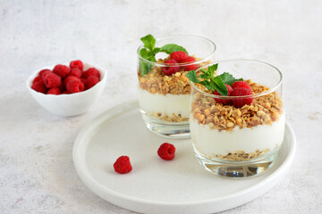 Two glasses with granola, yogurt, raspberry and mint leaves with bowl of raspberries