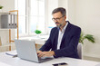 © Studio Romantic - Mature businessman, corporate worker or company director working in a modern office. Happy business man in a dark blue suit and glasses sitting at his white desk and typing on his laptop computer
