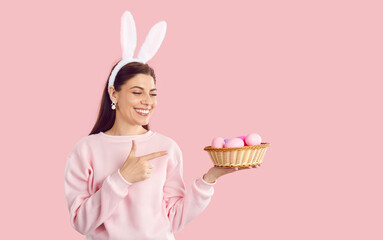 Happy beautiful woman wearing pink sweatshirt, earrings and bunny ears standing on pastel pink studio background, smiling and pointing at basket of pink Easter eggs that she is holding in her hand