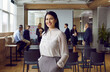© Studio Romantic - Portrait of young smiling brunette professional business coach woman in office looking at camera. Students, group of people on background. Independent, confident woman stands with hands in pockets.
