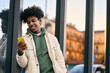 © insta_photos - Cool smiling young African American teen guy holding mobile phone standing at glass city wall. Happy stylish hipster teenager tech user using cell outdoors, looking at cellular checking smartphone.