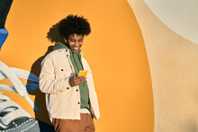 Happy Smiling Cool Gen Z Young African American Ethnic Stylish Hipster Guy Model Standing At Yellow City Urban Wall Using Cell Phone Mobile Device, Looking At Smartphone, Holding Cellphone.
