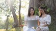 © Natee Meepian - Two Asian women are watching a video on a laptop together while enjoying a picnic in the park