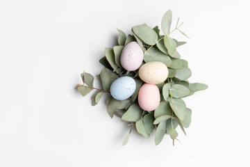 Wall Mural - Pastel coloured easter eggs, in a rustic nest made of eucalyptus leaves, on a white background, photographed from above.