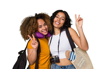 Two diverse friends on a beach vacation with vintage camera, beach bag, and travel pillow joyful and carefree showing a peace symbol with fingers.