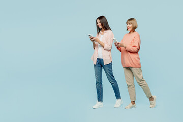 Full body smiling happy elder parent mom with young adult daughter two women together wear casual clothes hold in hand mobile cell phone walk go isolated on plain blue background. Family day concept.