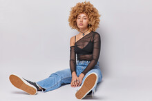 Photo Of Curly Haired Woman Has Serious Expression Wears Black Top Jeans And Sneakers Poses On Floor Against White Background. Lovely Teenage Girl In Fashionable Clothes Rests After Walking.