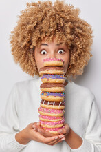 Photo Of Shocked Curly Haired Woman Holds Stack Of Delicious Appetizing Doughnuts Eats Harmful Food Stares Bugged Eyes Dressed In Casual Jumper Isolated Over White Background. Binge Eating Concept