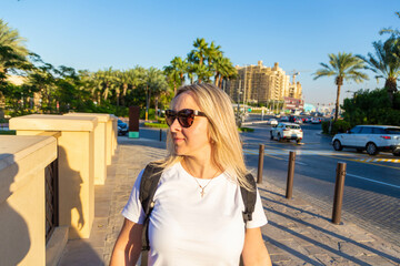 Wall Mural - Dubai resort area, a blonde tourist girl admires the stunning view of modern architece, bustling harbor, opulent boats, towering skyscrapers, and a bright sun with clouds while taking some selfies.