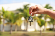 Real Estate Agent, Home Keys In Female Hand On Background Of House Surrounded By Palm Trees. Buying Or Rental A Villa On Ocean Coast, Removal To Tropical Country