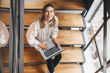 Beautiful optimistic young woman wear knitting vest and white shirt sitting on wood stairs indoors at home while using laptop computer. View from above. Girl smiling and look at camera.