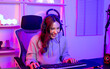 © Ann Rodchua - Beautiful happy young female game online streamer or caster smiling with fun, succeed for winner competition, using computer and keyboard to play, sitting in room at home with decorated neon light.