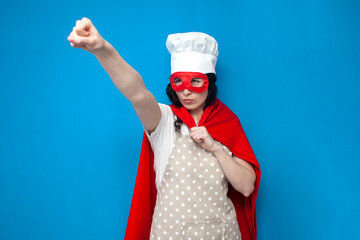 girl chef in superman costume holds kitchen item on blue background, woman housewife in superhero mask points forward