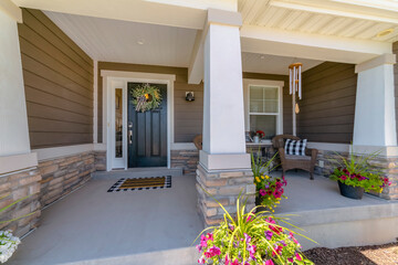 Sticker - Facade of a house with brown wood lap siding and decorative porch. Black front door with wreath and sidelight near the woven chairs with potted flowers on the right.